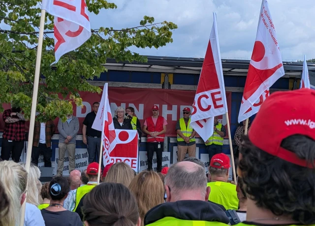 Eine Frau spricht auf einer Bühne hinter einem Rednerpult. Hinter ihr stehen weitere Personen. Im Vordergrund sind Menschen mit Flaggen zu sehen, die der Vortragenden zuhören.