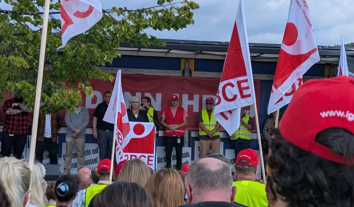 Eine Frau spricht auf einer Bühne hinter einem Rednerpult. Hinter ihr stehen weitere Personen. Im Vordergrund sind Menschen mit Flaggen zu sehen, die der Vortragenden zuhören.