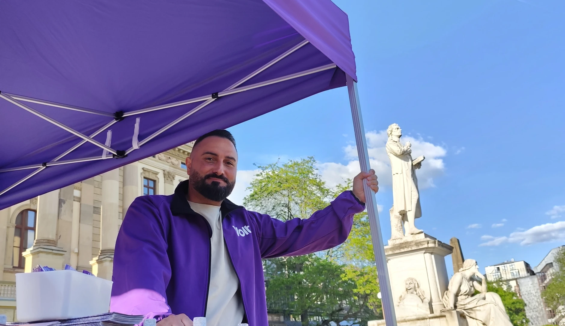 Ein Mann steht an einem Infostand. Im Hintergrund ist eine Statue zu sehen.