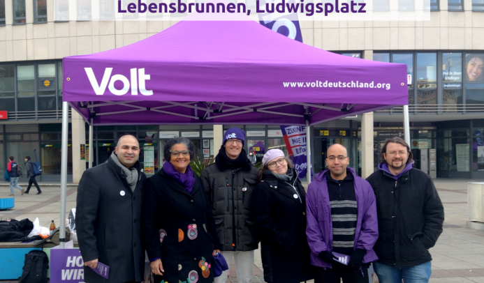Auf dem Foto sieht man einen Infostand von Volt MaLu mit Pavillon und von links nach rechts Burak, Beate, Tommy, Michaela, Johannes, Michael. Sie stehen am Berliner Platz in Ludwigshafen. Der Text bewirbt den Infostand am 6.9. am Lebensbrunnen, Ludwigshafen.