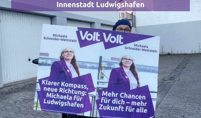 Zu sehen ist Michaela, wie sie hinter einem Wahlplakat hervorlugt. Das Plakat ist an einem Bollerwagen befestigt. Es ist das Plakat mit den Slogans