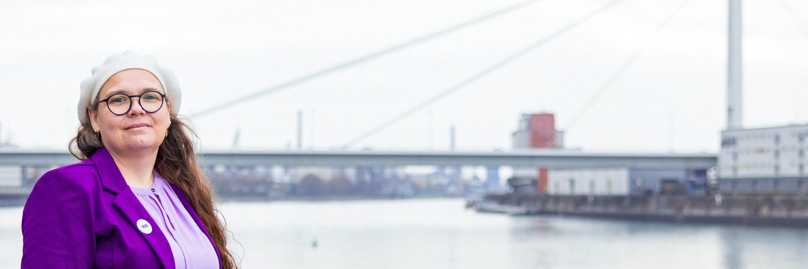 Portrait von Michaela am Rheinufer mit kühnem Blick und der Pylonbrücke im Hintergrund.