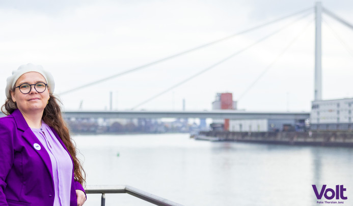 Portrait von Michaela am Rheinufer mit kühnem Blick und der Pylonbrücke im Hintergrund.