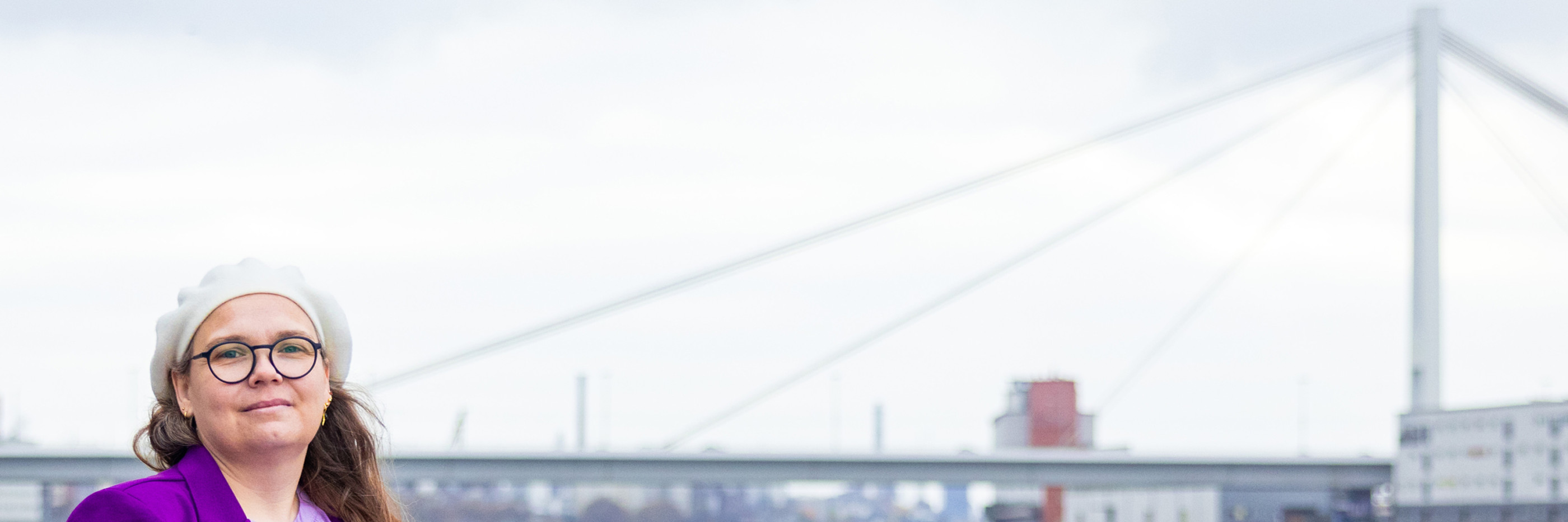 Portrait von Michaela am Rheinufer mit kühnem Blick und der Pylonbrücke im Hintergrund.