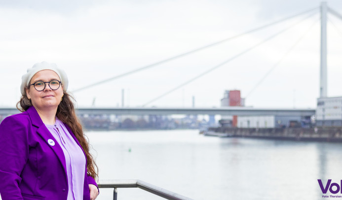 Portrait von Michaela am Rheinufer mit kühnem Blick und der Pylonbrücke im Hintergrund.
