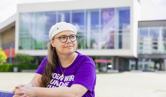 Auf dem Foto sieht man Michaela vor dem Pfalzbau. Sie stützt lässig ihre Arme auf einem Geländer ab und schaut mit zuversichtlichem und hoffnungsvollem Blick an der Kamera vorbei in die Weite. Foto: Thorsten Janz.