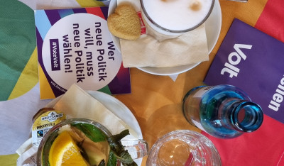 Tisch von oben mit diversen Heißgetränken, einer Volt Flagge in Regenbogen und diversen Volt Flyern