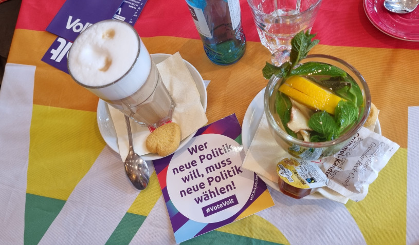 Latte macchiatto und Minztee auf einer regenbogenfarbigen Volt Flagge und Aufkleber von Volt