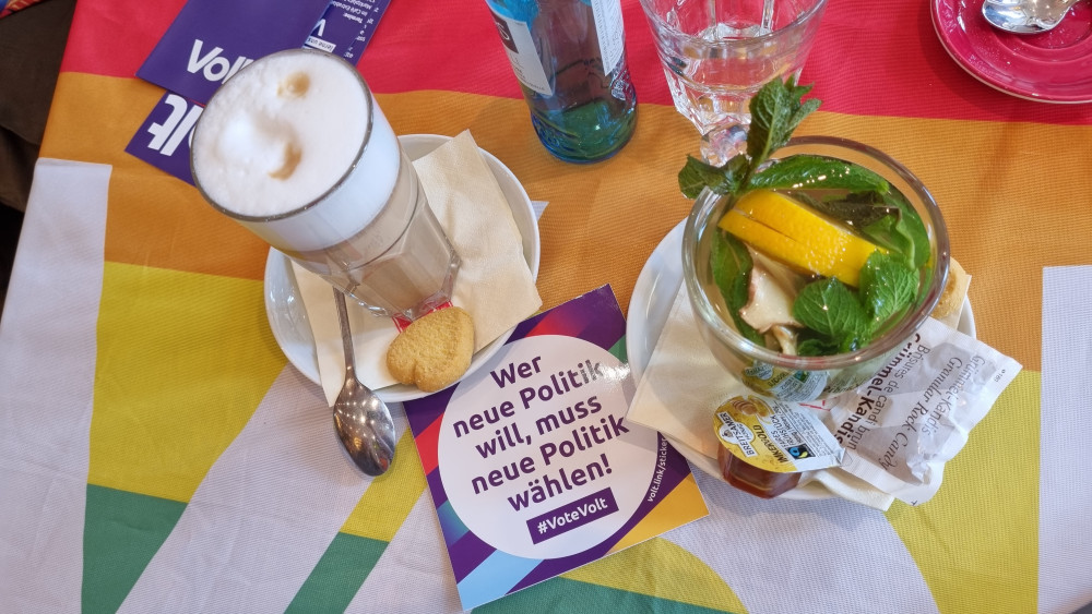 Latte macchiatto und Minztee auf einer regenbogenfarbigen Volt Flagge und Aufkleber von Volt