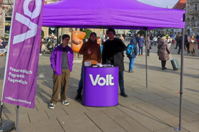 drei Mensch(Volta) hinter einem tisch mit Volt logo unter einem lila Pavillon links daneben eine Voltflagge