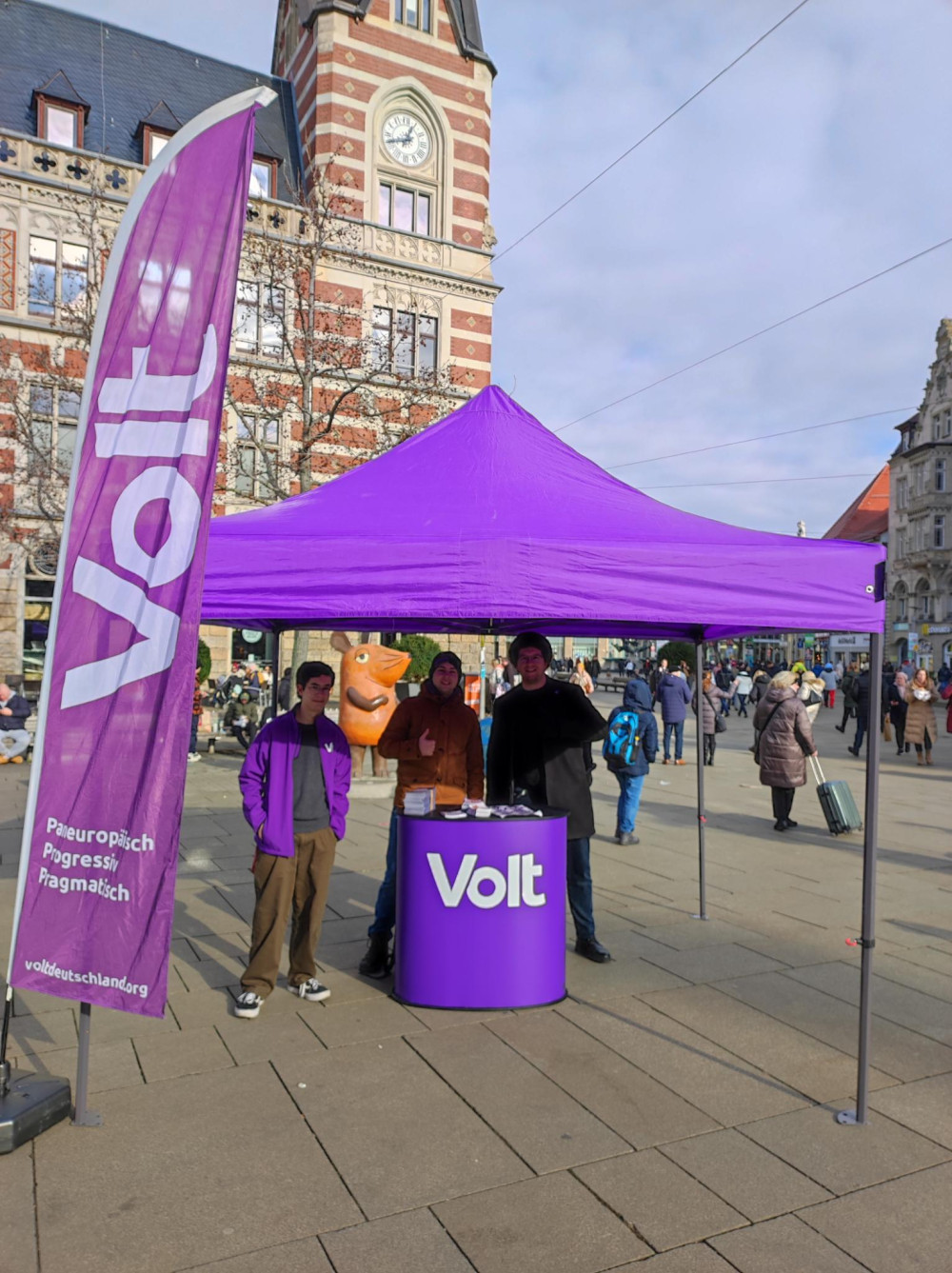 drei Mensch(Volta) hinter einem tisch mit Volt logo unter einem lila Pavillon links daneben eine Voltflagge