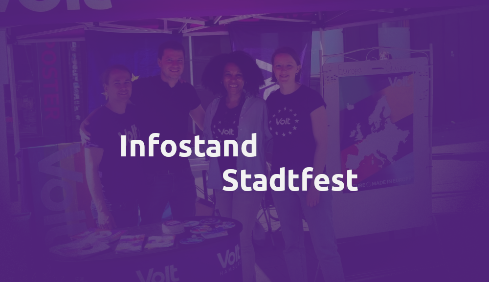 Durch eine Volt violette Maske sehen wir einen Infostand von Volt mit vier nett lächelnden Leuten im am Infostand, einer Regenbogenflagge von Volt und einem Tisch mit verschiedenen Flyern und Stickern. Im Vordergrund steht geschrieben: Infostand Stadtfest